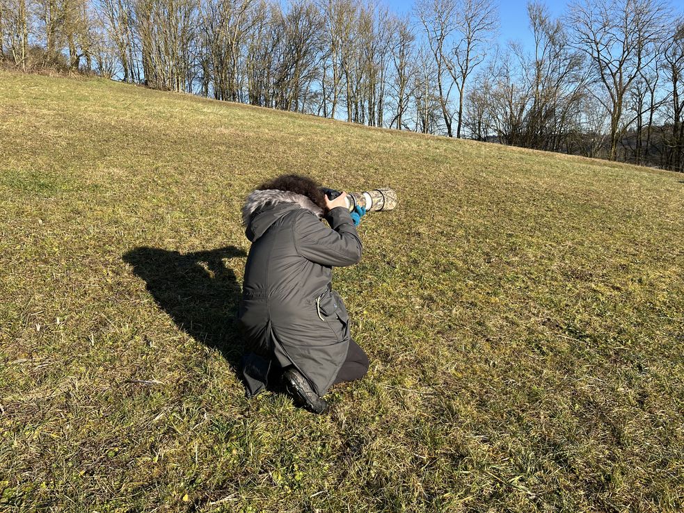 Frau kniet auf der Wiese, in der Hand ein Fotoapparat mit Teleobjektiv